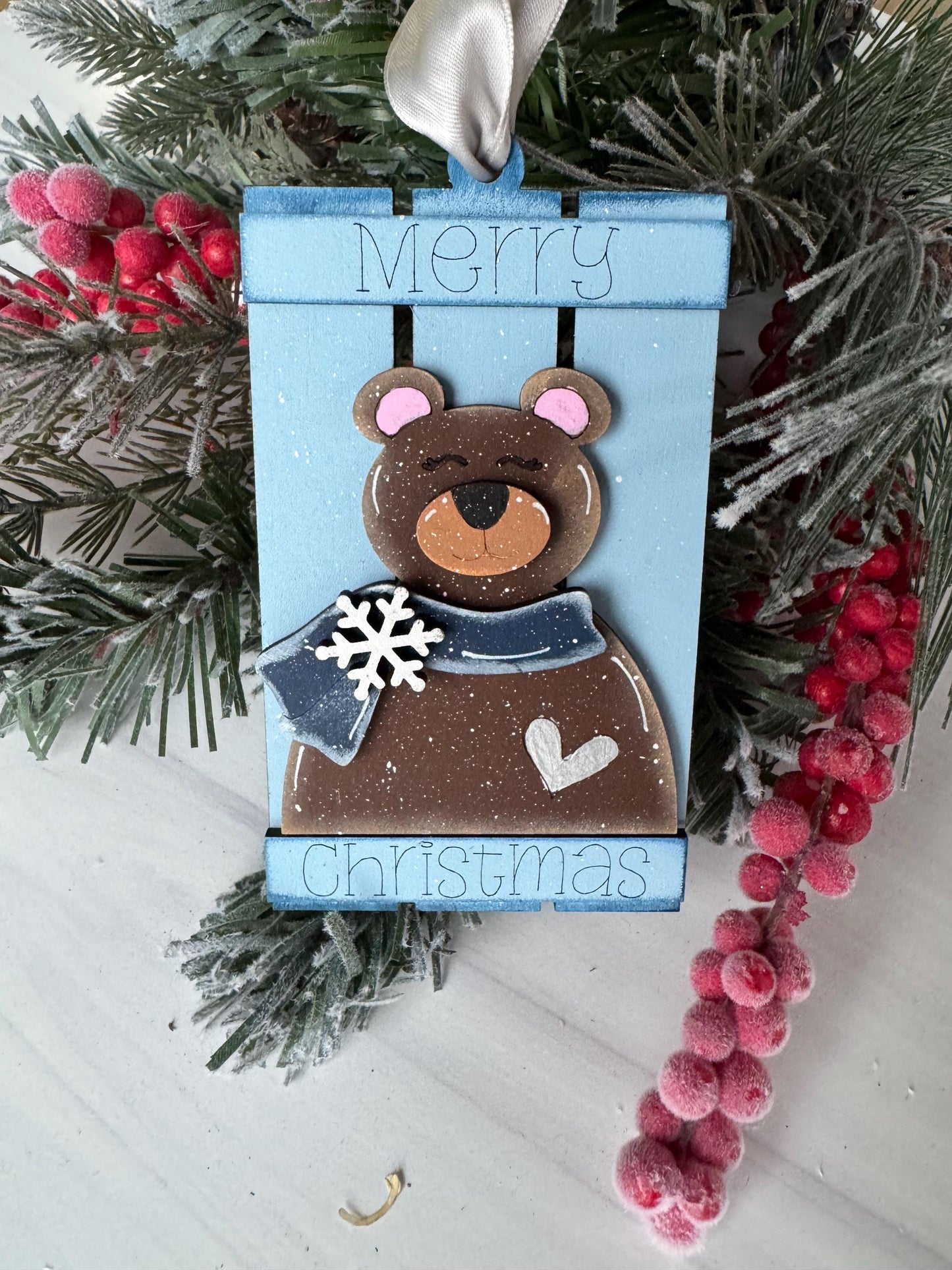 A blue Christmas ornament with a bear and snowflakes, placed on a snowy surface with red berries and pine branches.