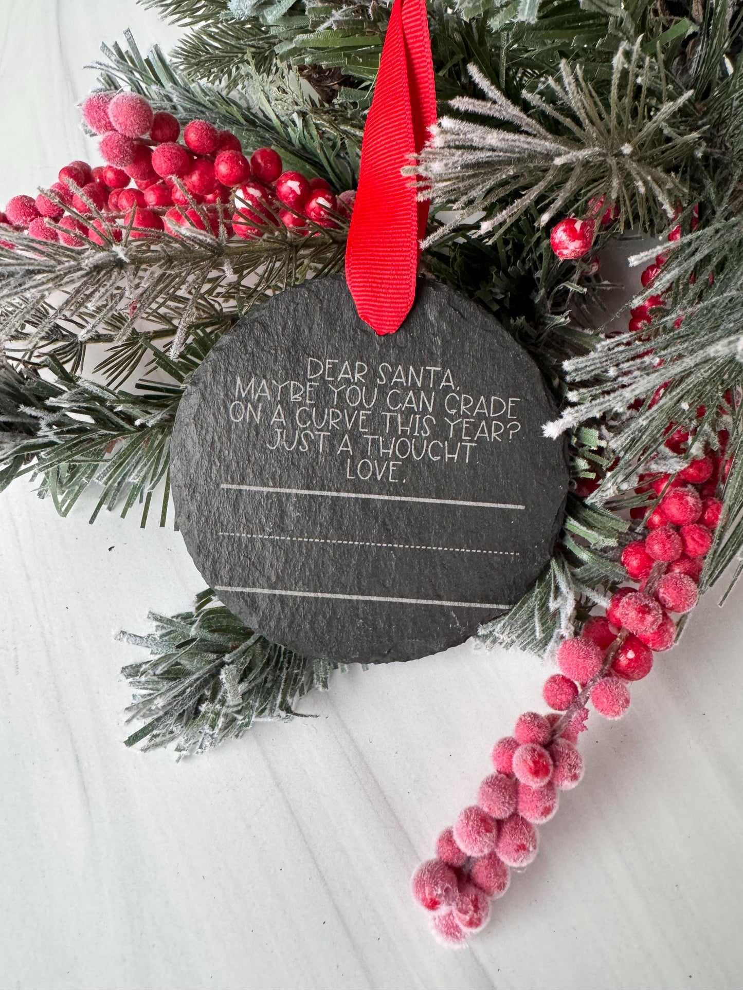A black slate with a message written on it, surrounded by a wreath of pine branches and red berries, placed on a white surface.