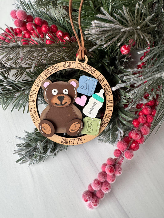 A wooden ornament with a teddy bear and various baby-related items, such as a bottle and a pacifier, is displayed on a pine branch with red berries.