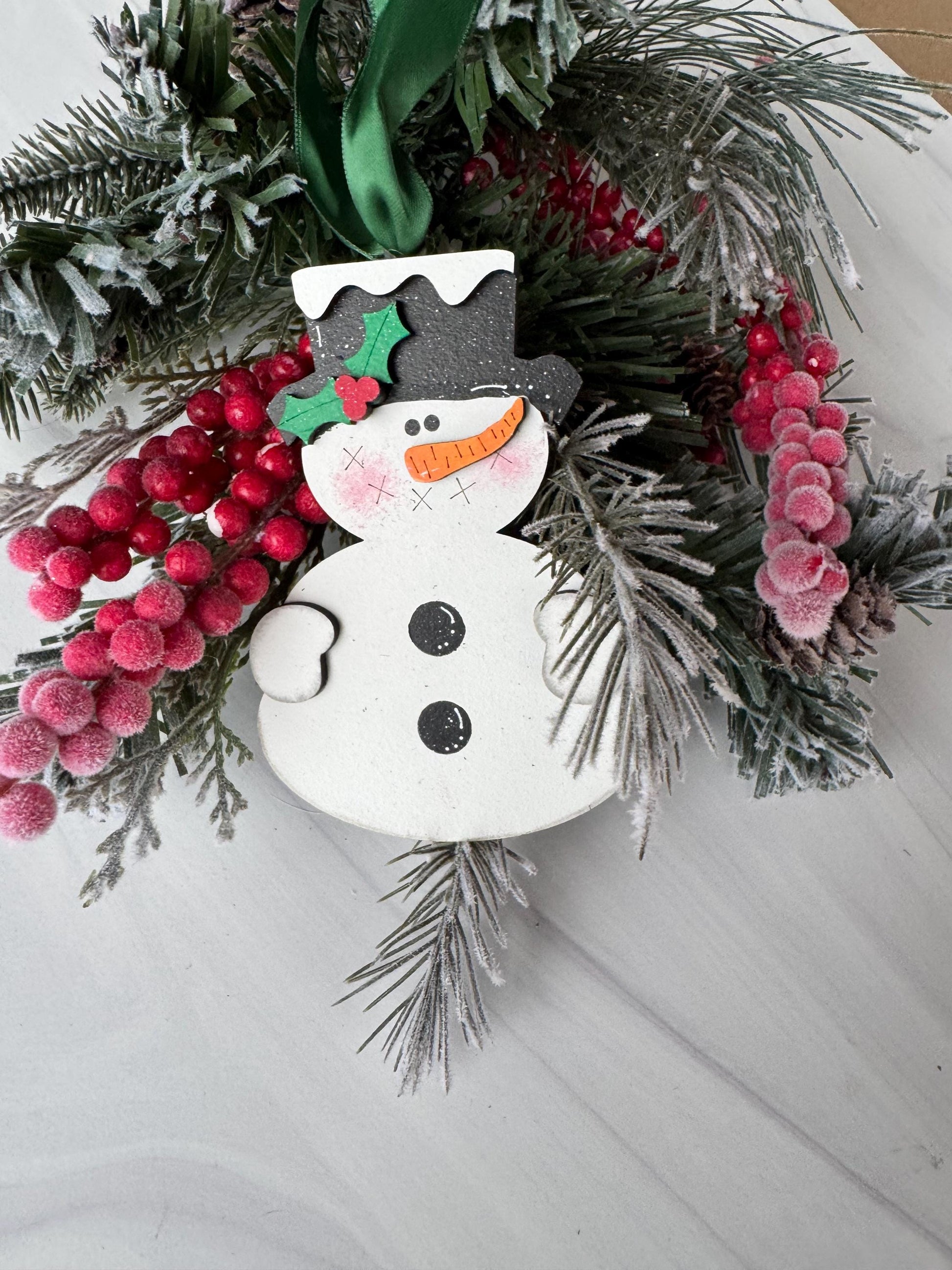 A festive wreath adorned with red berries, pine branches, and a snowman ornament is displayed on a white surface.