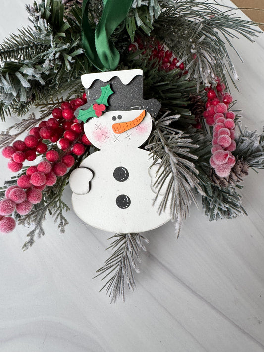 A festive wreath adorned with red berries, pine branches, and a snowman ornament is displayed on a white surface.