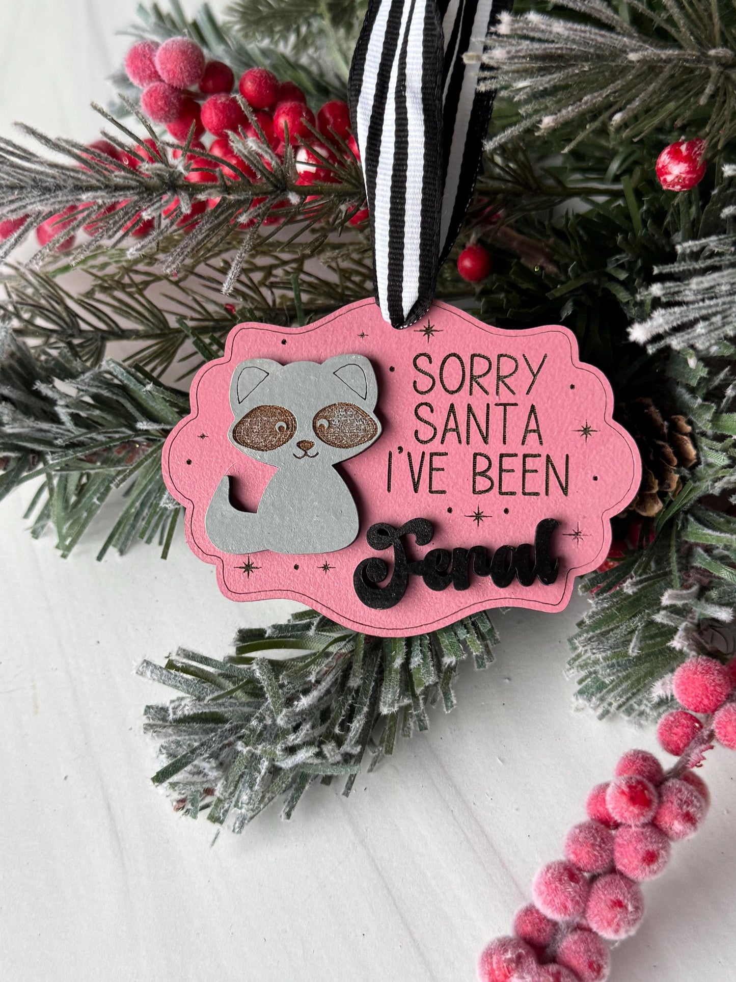 A pink ornament with a cat design and the text "Sorry Santa I've been away" is displayed among pine branches and red berries.