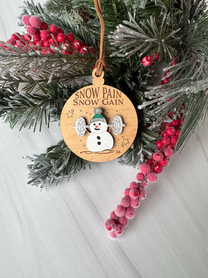 A wooden ornament with a snowman design and the text "Snow Pain Gain" is placed on a pine branch with red berries.