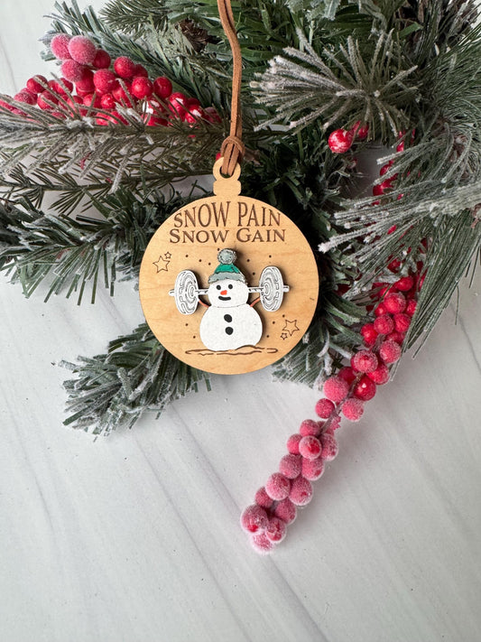 A wooden ornament with a snowman design and the text "Snow Pain Gain" is placed on a pine branch with red berries.