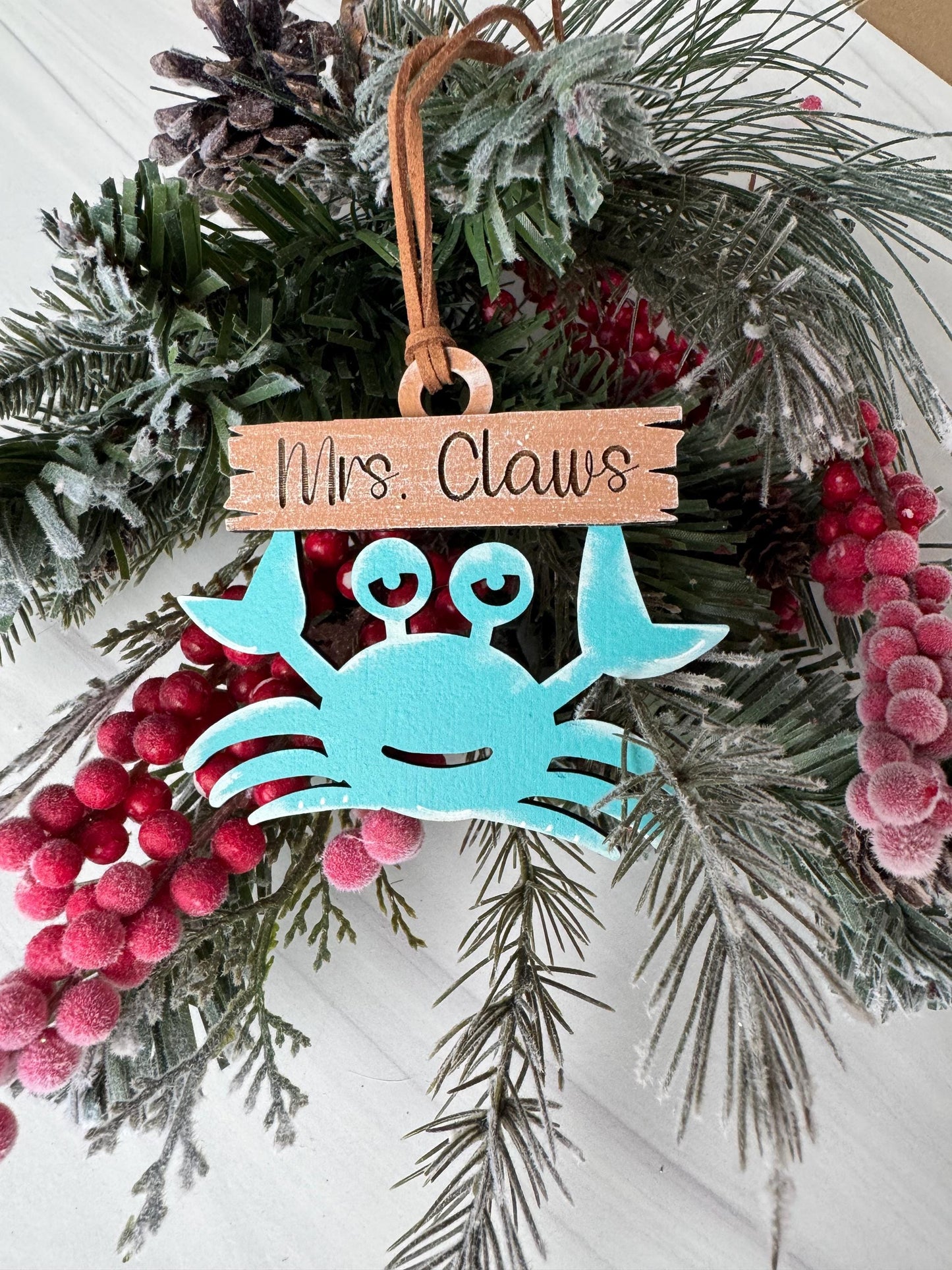 A blue crab ornament is displayed on a wreath of pine branches and berries, with a wooden sign attached to it.