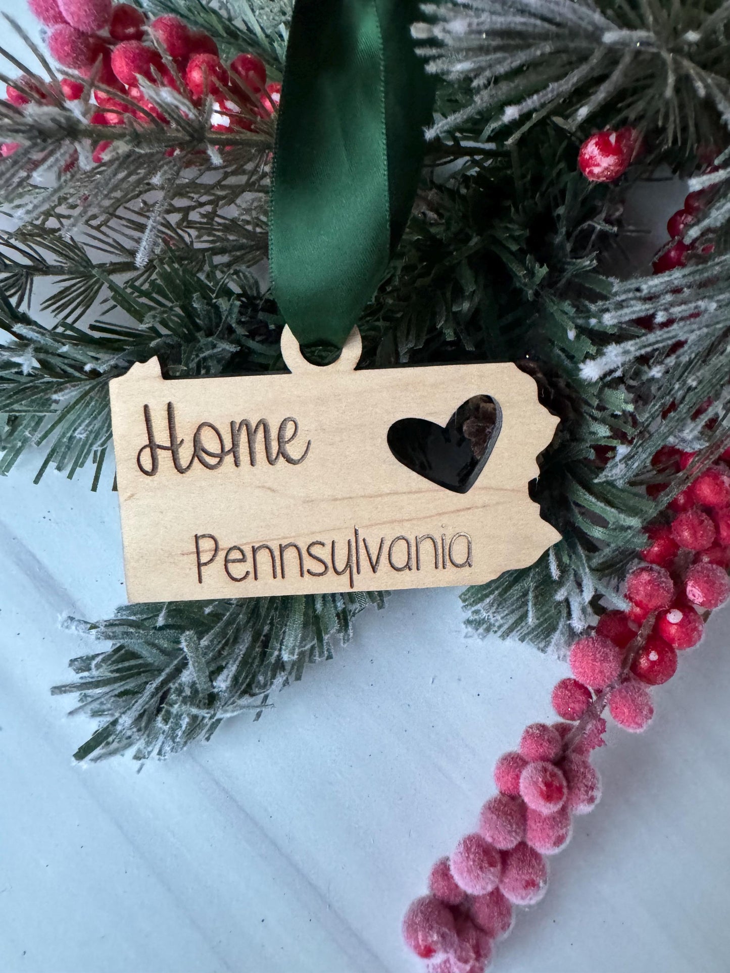 A wooden ornament shaped like the state of Pennsylvania, with the word &quot;Home&quot; written on it, is hanging on a Christmas tree branch with red berries.