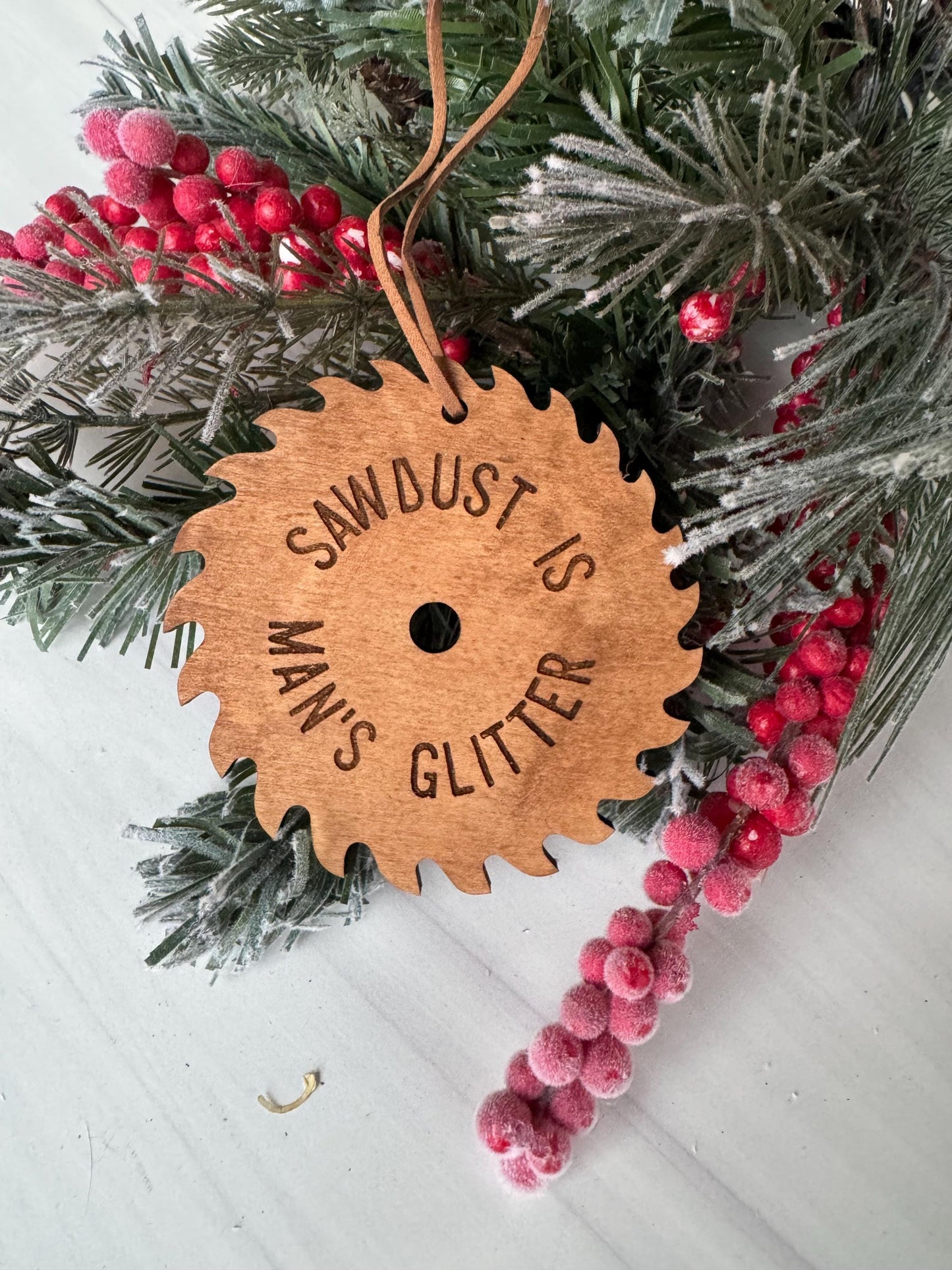 A wooden ornament with the text &quot;SAWBOARD IS GLITTER&quot; engraved on it, surrounded by a sprig of pine needles and red berries.
