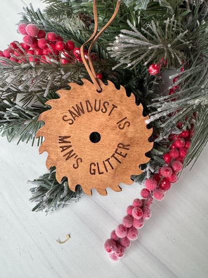 A wooden ornament with the text &quot;SAWBOARD IS GLITTER&quot; engraved on it, surrounded by a sprig of pine needles and red berries.