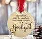 A wooden ornament with a red ribbon hanging from it, featuring a humorous text that reads "My books may be naughty but Santa thinks I'm a good girl".