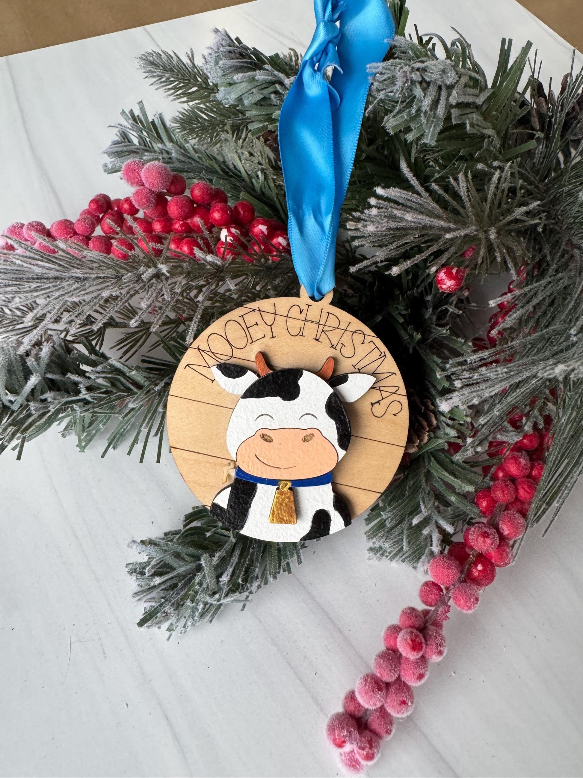 A Christmas ornament featuring a cow is displayed on a white surface, surrounded by a wreath of pine branches and red berries.