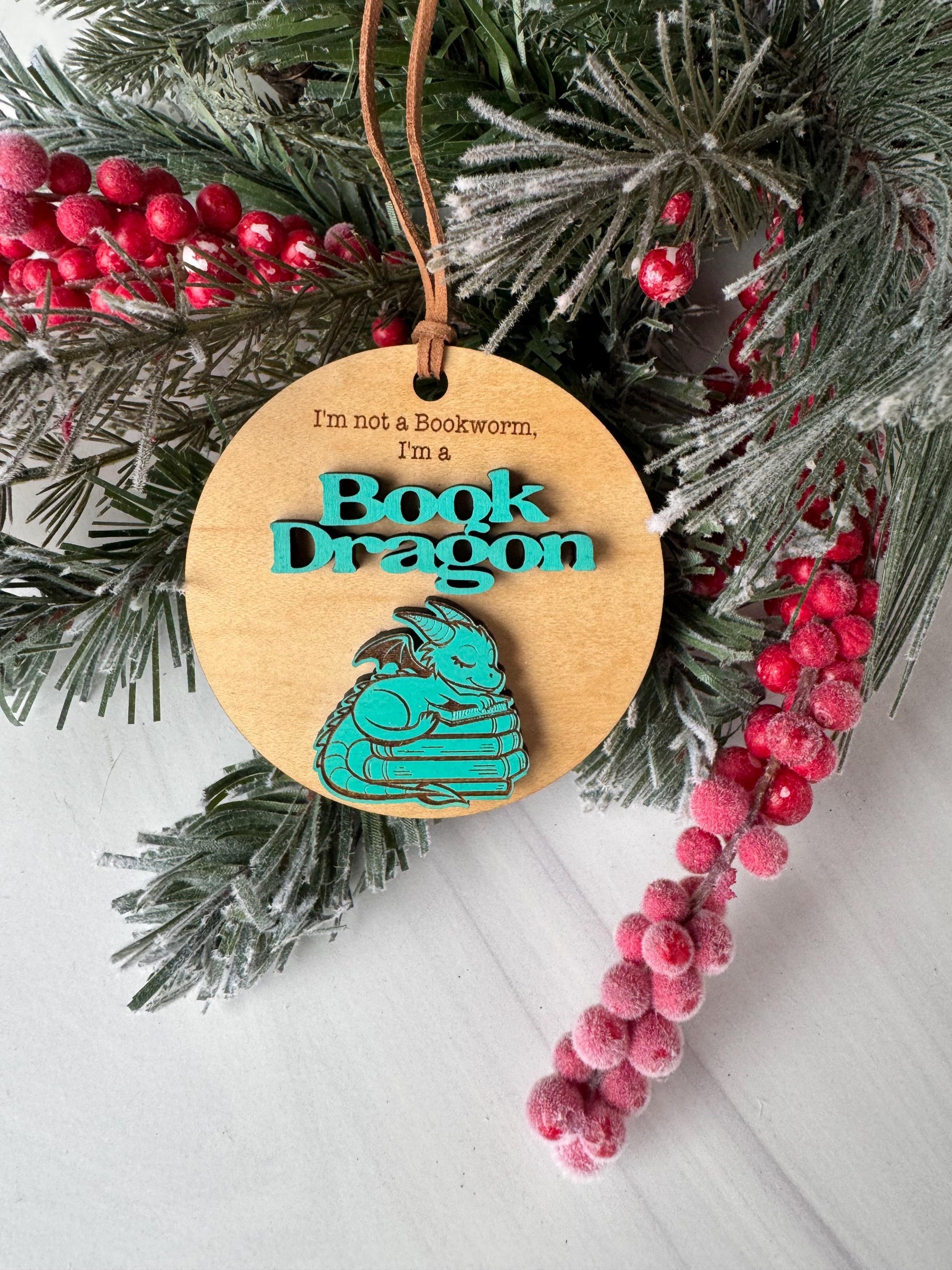 A wooden ornament with a dragon design, surrounded by a wreath of pine branches and red berries, placed on a white surface.