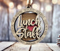 A wooden ornament with the word "Lunch Staff" written on it, surrounded by the words "Hard working - Dedicated - Generous - Good with kids - Consistent - Effortless - Flexible" in a circular frame.