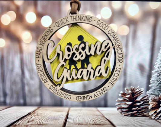A wooden ornament with the words "Crossing Guard" written on it, surrounded by a circular frame. The ornament is placed on a wooden surface, with pine cones visible in the background.