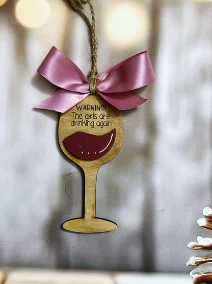 A wooden ornament shaped like a wine glass with a pink ribbon and a warning message hanging from it.