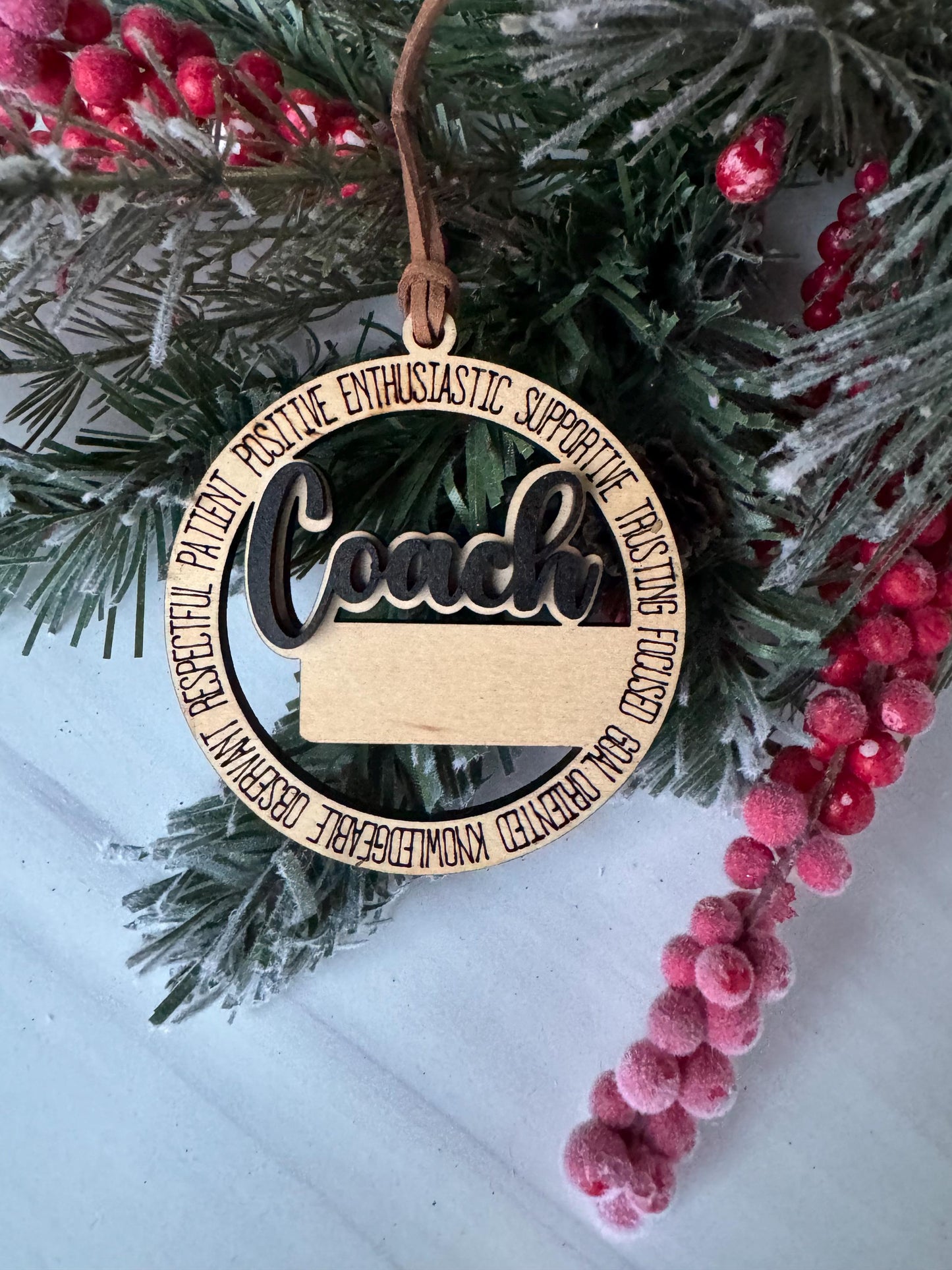 A wooden ornament with the word &quot;Coach&quot; on it, surrounded by pine branches and red berries, placed on a white surface.