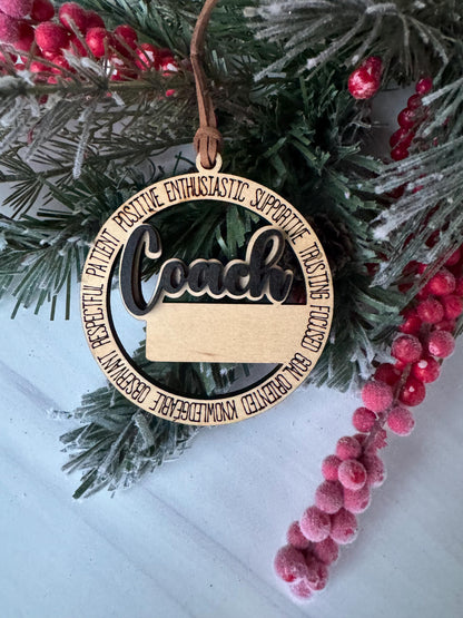 A wooden ornament with the word &quot;Coach&quot; on it, surrounded by pine branches and red berries, placed on a white surface.