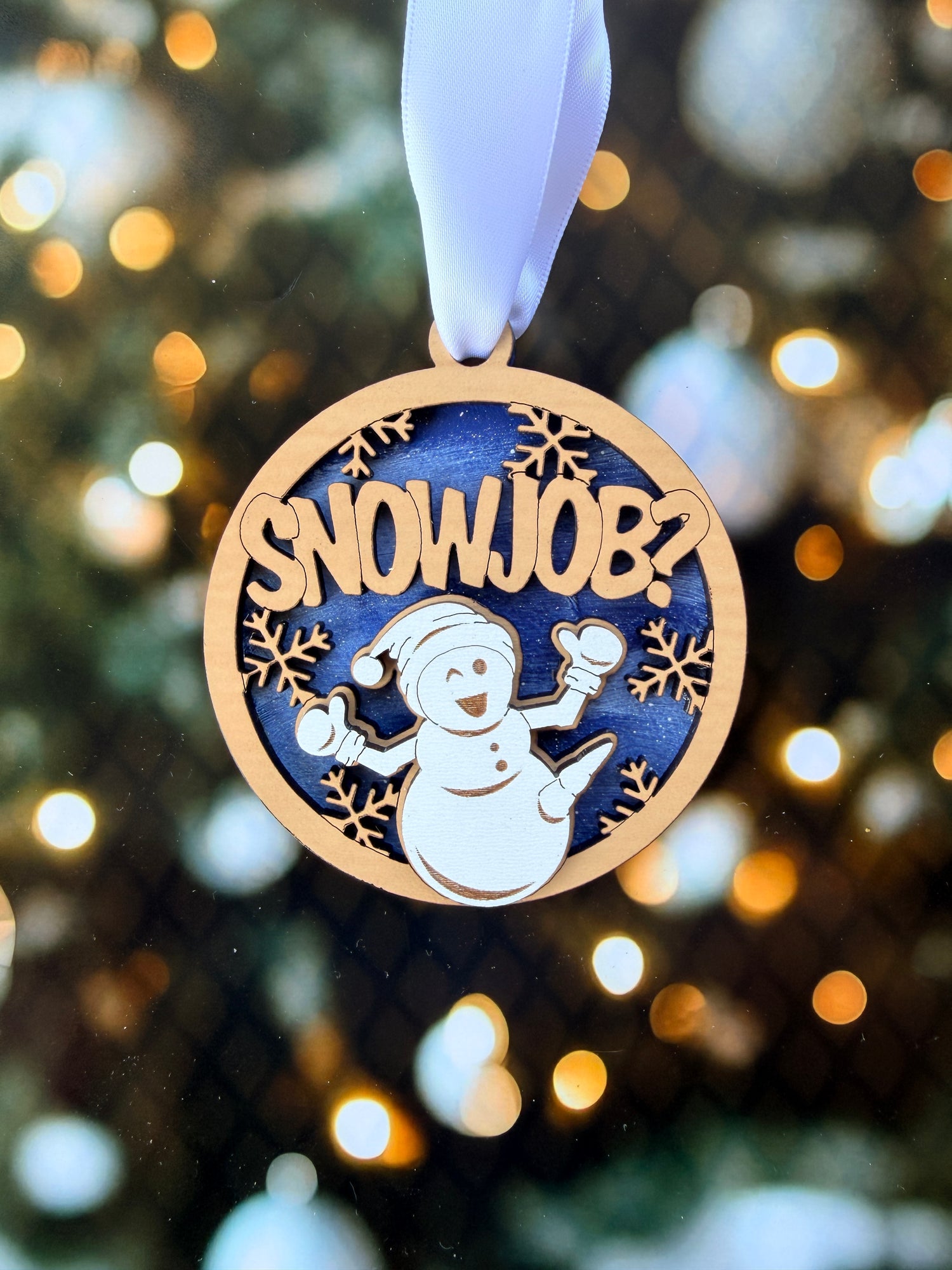 A wooden ornament with a snowman design and the text "SNOWJOB?" is displayed against a blurred background with twinkling lights.