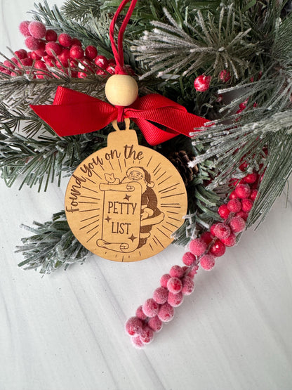 A wooden ornament with a red ribbon and the text "The list" is surrounded by a wreath of pine branches and red berries on a white surface.