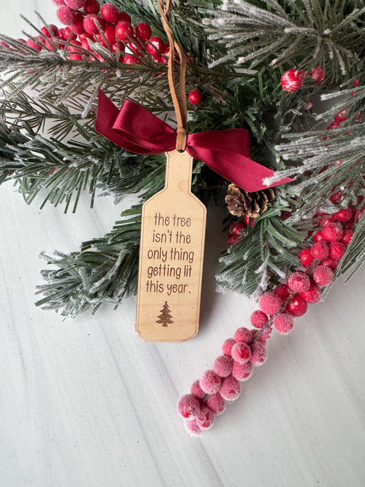 A wooden tag with a Christmas tree decoration attached to it, surrounded by pine branches, red berries, and a red ribbon.