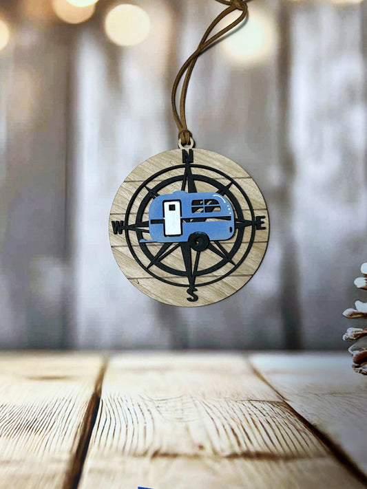 A wooden pendant with a compass design and a blue van or camper on it, hanging on a string against a blurred background.
