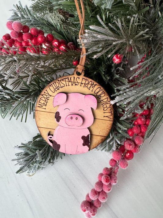 A Christmas ornament featuring a pink pig is displayed on a pine branch with red berries.