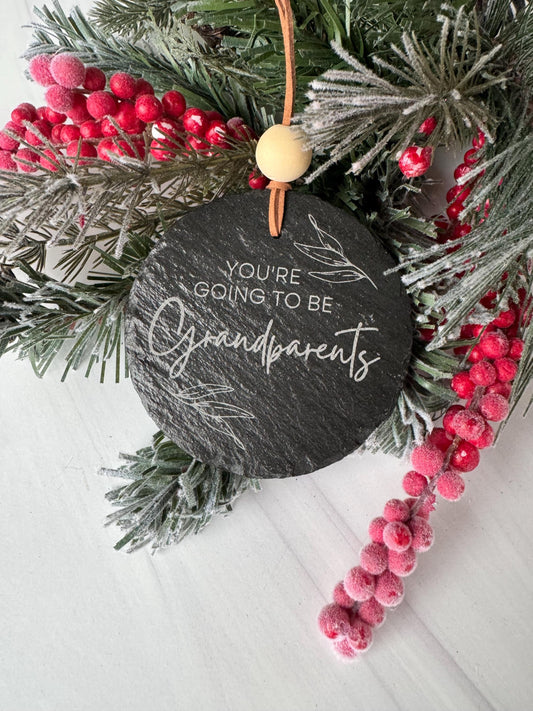 A black slate ornament with the text &quot;You&#39;re going to be grandparents&quot; is surrounded by a wreath of pine branches and red berries on a white surface.