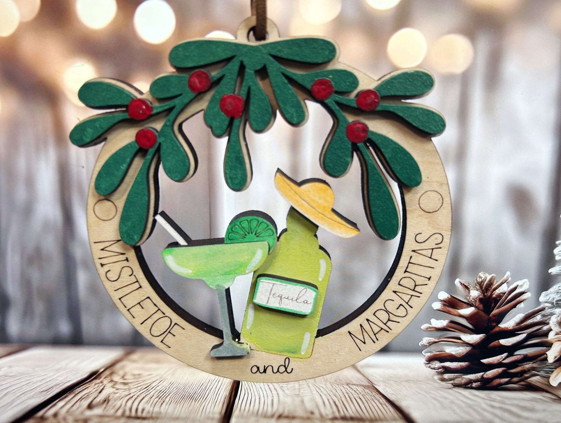 A wooden ornament with a festive design, featuring a green mistletoe, a tequila bottle, and a margarita glass, surrounded by the words "Mistletoe" and " margaritas".