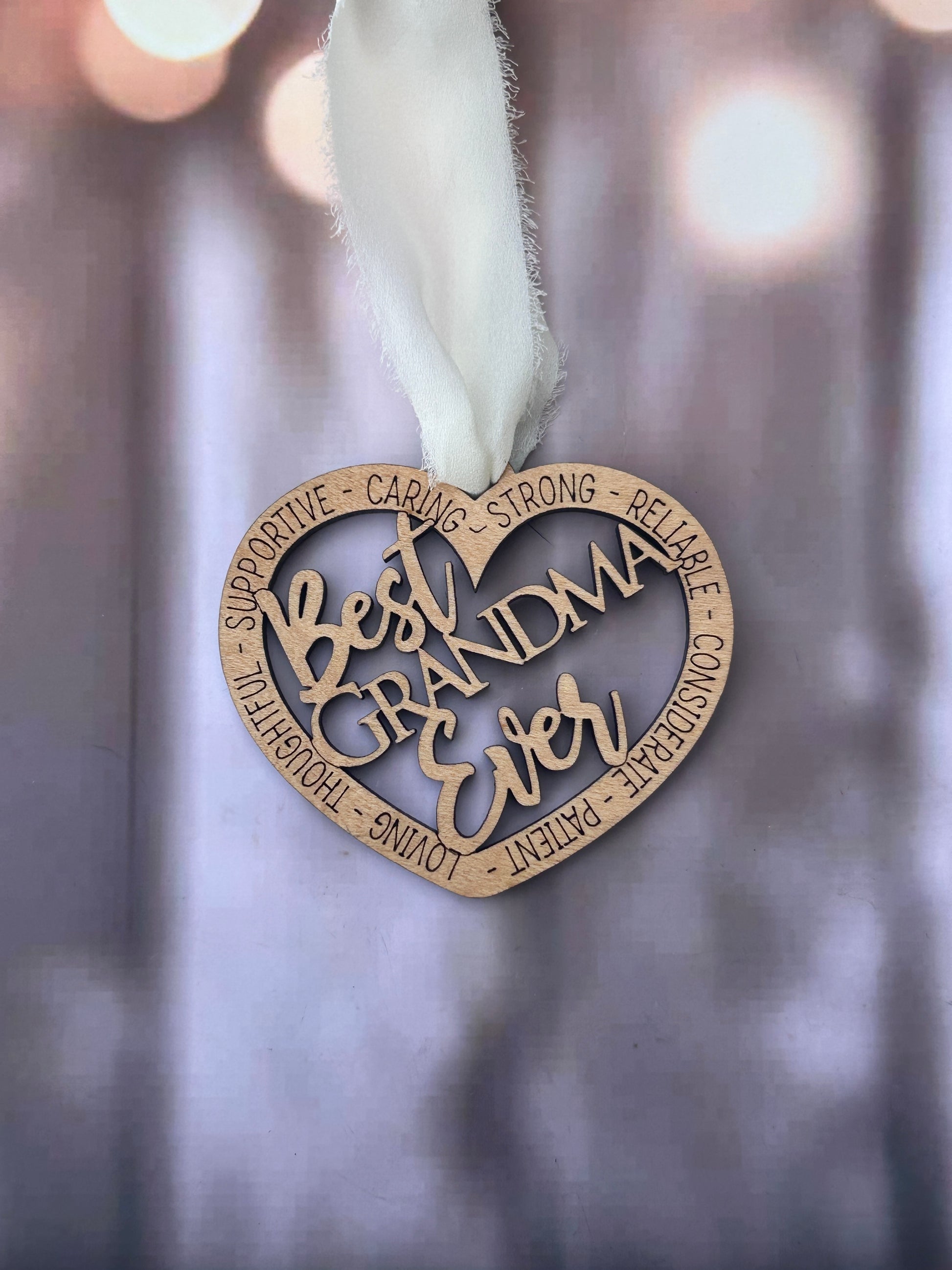 A heart-shaped ornament with the words "Best Grandma Ever" written on it, hanging against a blurred background.