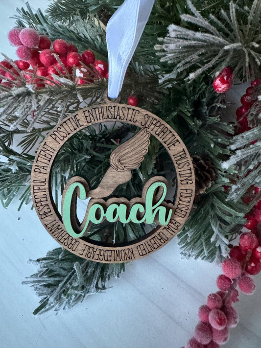 A wooden ornament with the word "Coach" written on it, hanging on a Christmas tree with red berries and pine branches.