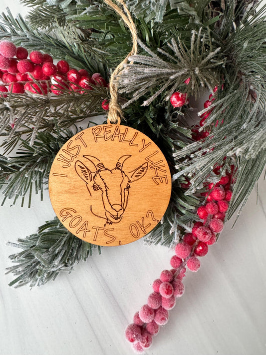 A wooden ornament with a goat design, surrounded by pine branches and red berries, with the text &quot;I really like goats&quot; engraved on it.