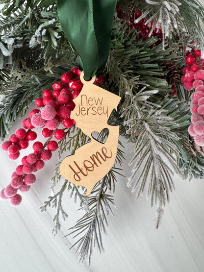 A wooden ornament shaped like the state of New Jersey, with the word "New Jersey" written on it, is hanging from a pine branch with red berries.