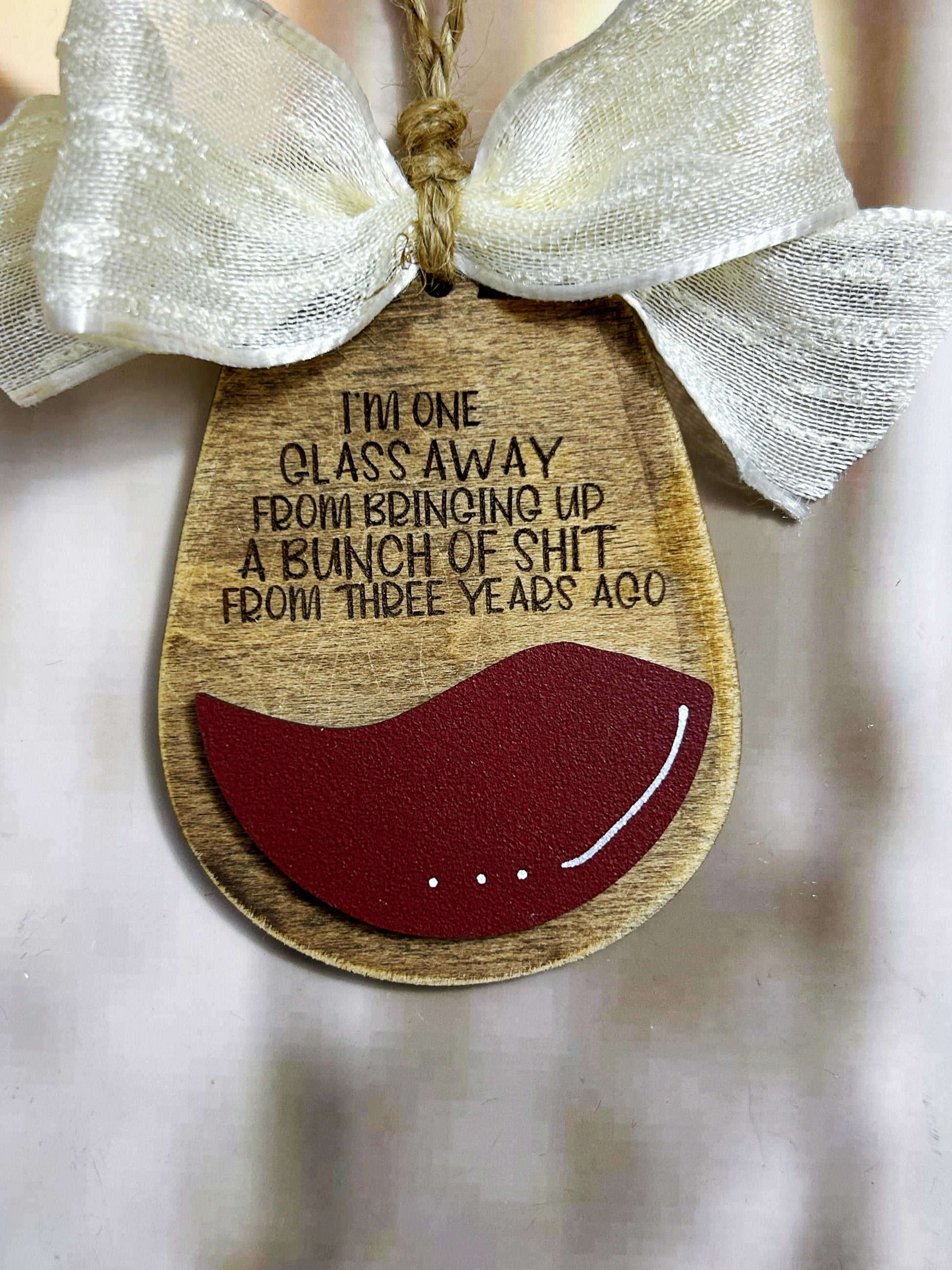 A wooden ornament with a red wine glass and a white ribbon, featuring a humorous message about a glass being taken away from a bunch of shit from three years ago.