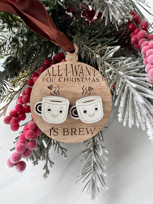 A wooden ornament with a wreath of pine branches and red berries, featuring a pair of cartoon coffee cups and the text &quot;All I want for Christmas is coffee&quot;.