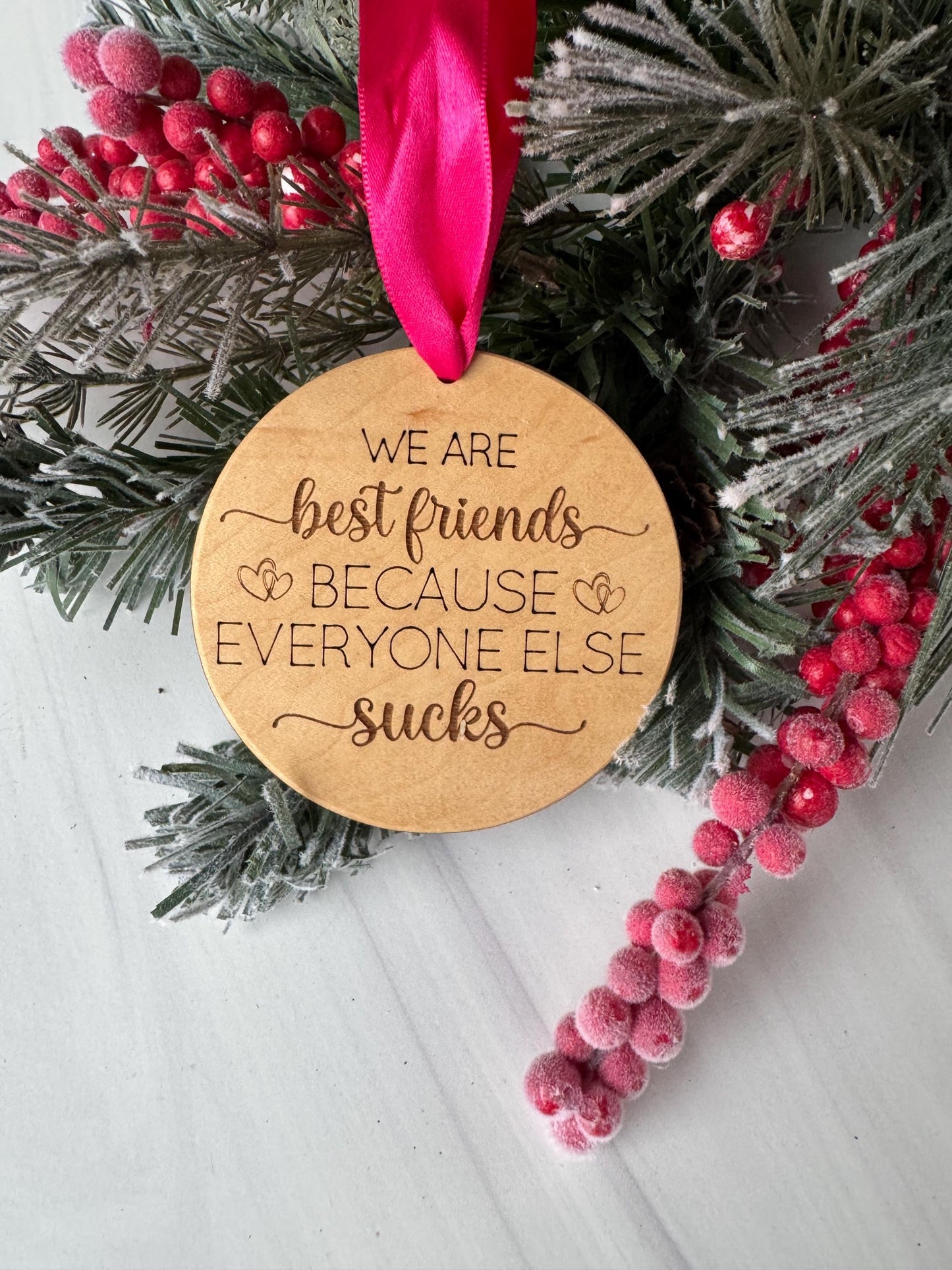 A wooden ornament with a ribbon, surrounded by pine branches and red berries, with the text &quot;WE ARE BEST FRIENDS EVERYONE ELSE SUCKS&quot; engraved on it.
