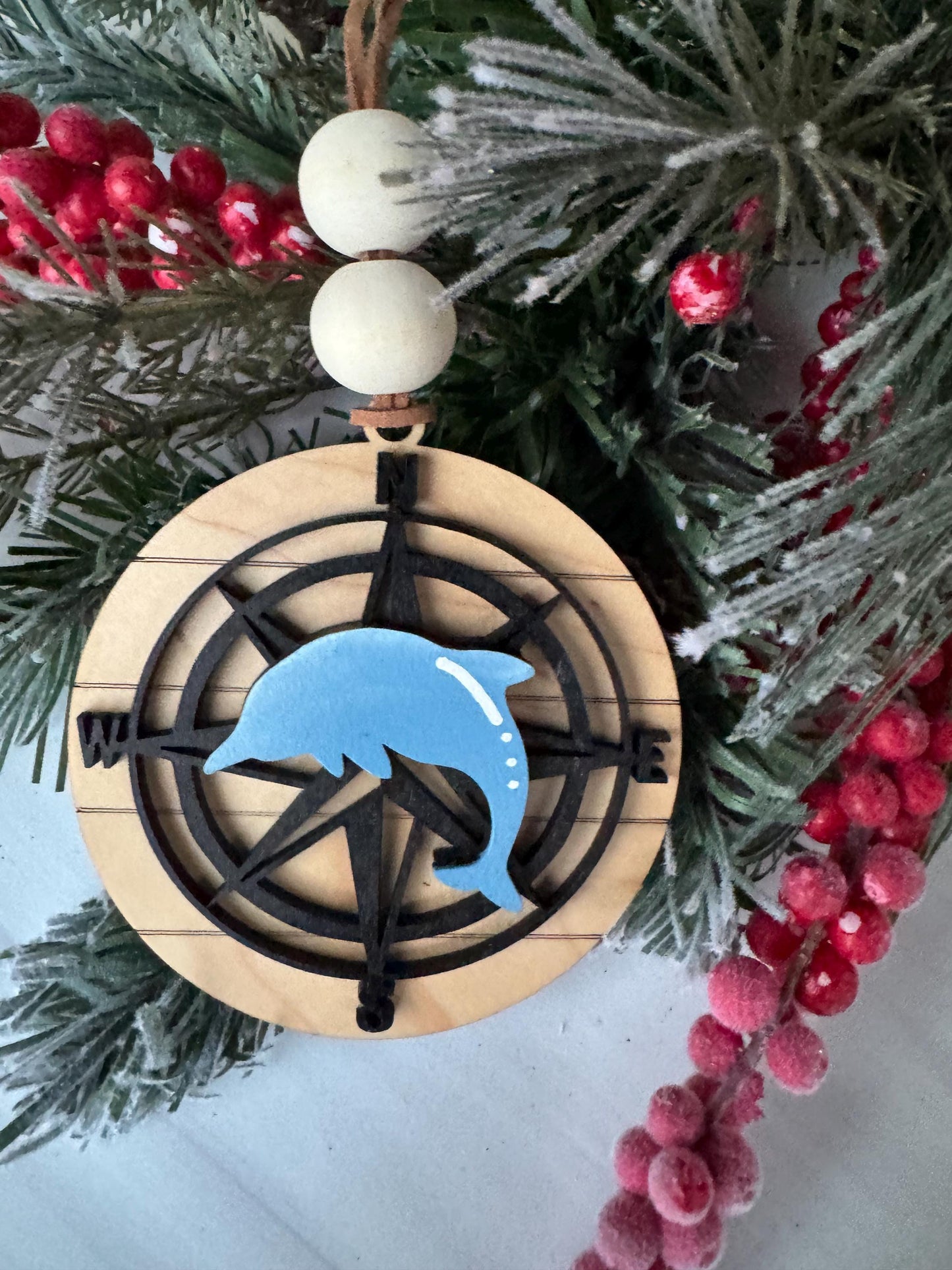 A wooden ornament with a compass design and a blue dolphin figure is displayed on a Christmas tree, surrounded by red berries and pine branches.