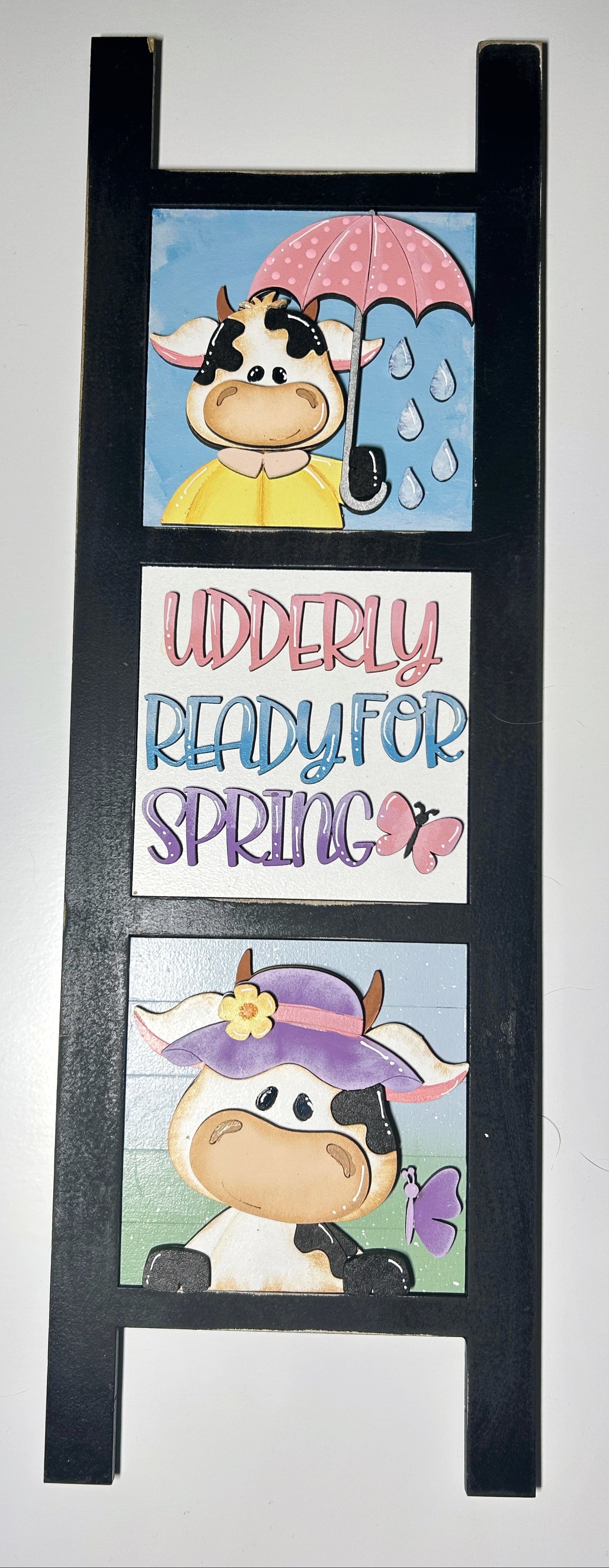 Decorative ladder with cow illustrations and 'Udderly Ready for Spring' text on a white background

