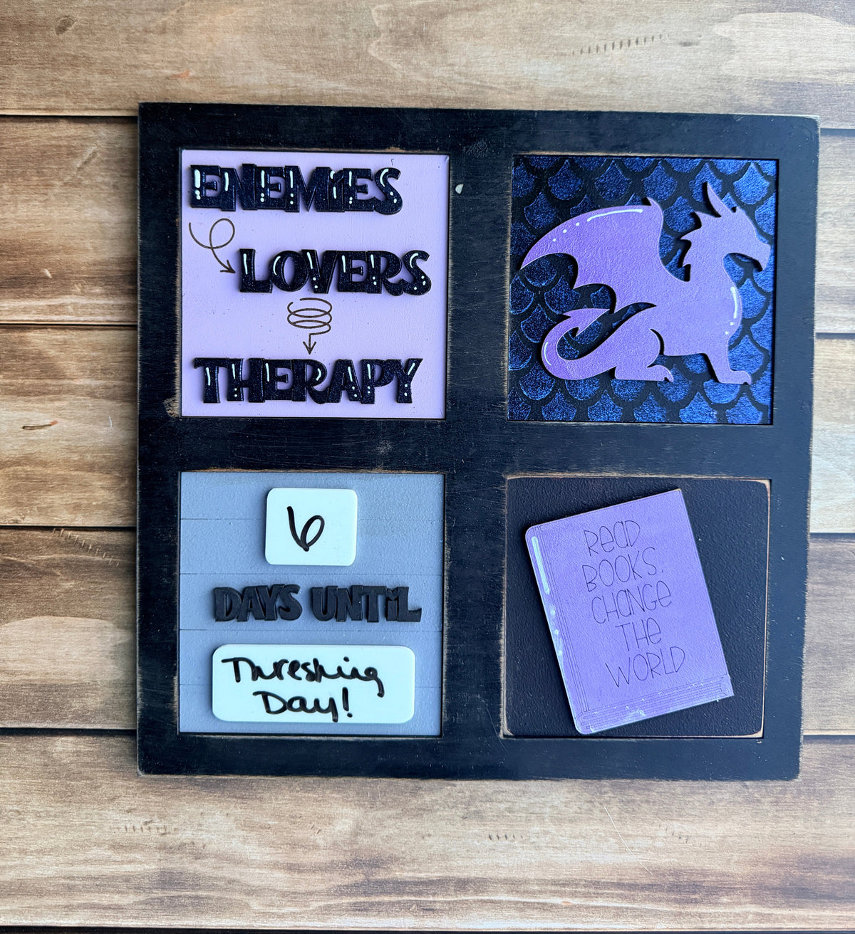 A black square decorative board or tray with four quadrants, photographed on a wooden surface. The top-left section contains a small purple card reading 'I love you so much'; the top-right has a white note and black lettering; the bottom-left shows a purple ghost shape over a dark patterned background; the bottom-right has a lavender card with black text and a small doodle.