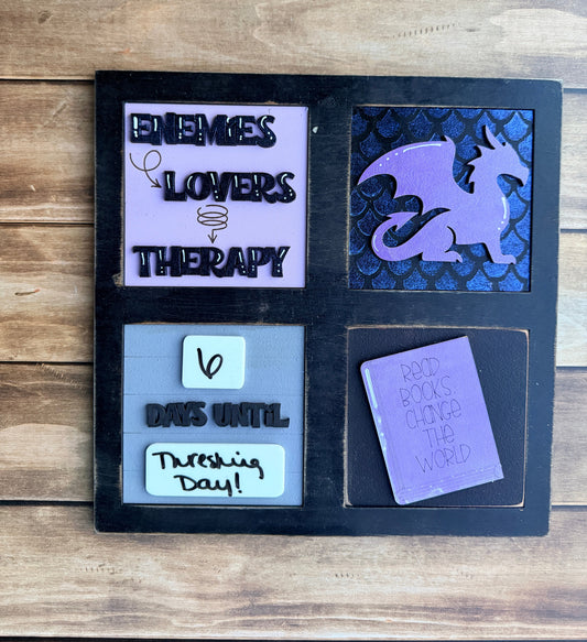 A black square decorative board or tray with four quadrants, photographed on a wooden surface. The top-left section contains a small purple card reading 'I love you so much'; the top-right has a white note and black lettering; the bottom-left shows a purple ghost shape over a dark patterned background; the bottom-right has a lavender card with black text and a small doodle.