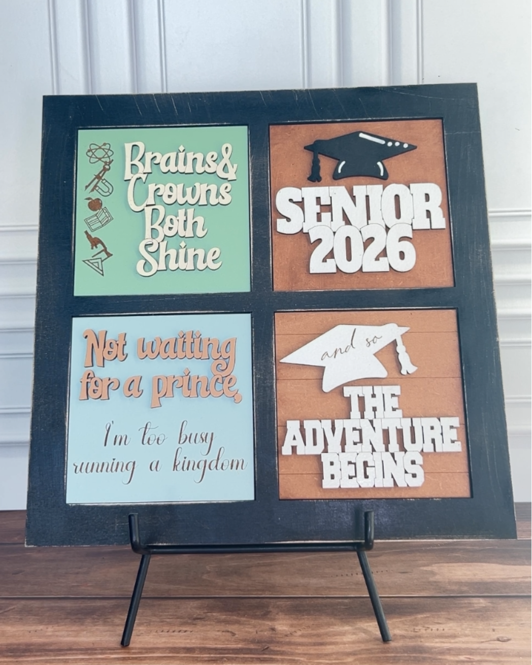 Decorative sign with graduation-themed sayings on a stand against a white paneled wall.