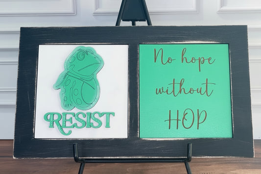 Two signs on a stand with a frog graphic and text, against a white paneled wall.