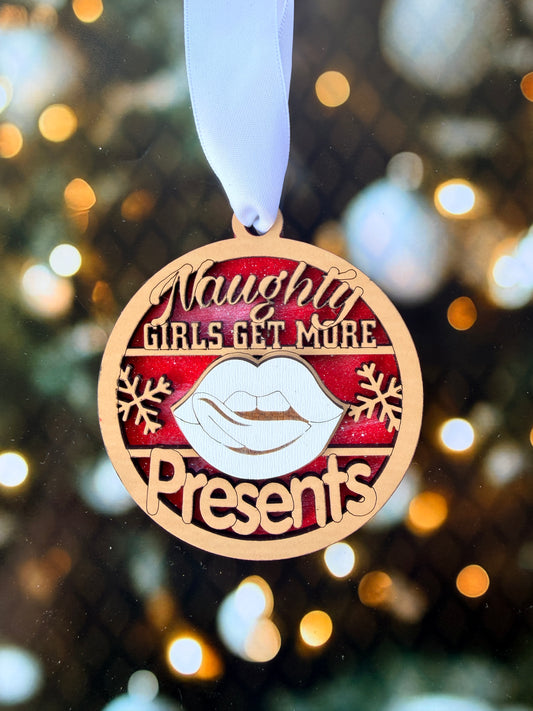 A close-up of a round ornament with a ribbon, featuring the text "Naughty Girls Get More" and a snowflake design.