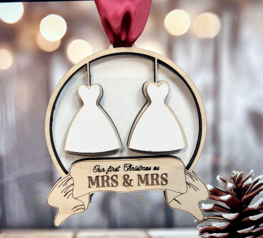 A wooden ornament with two white dress silhouettes hanging from a red ribbon, and a banner below that reads "Our first Christmas as MRS & MRS".