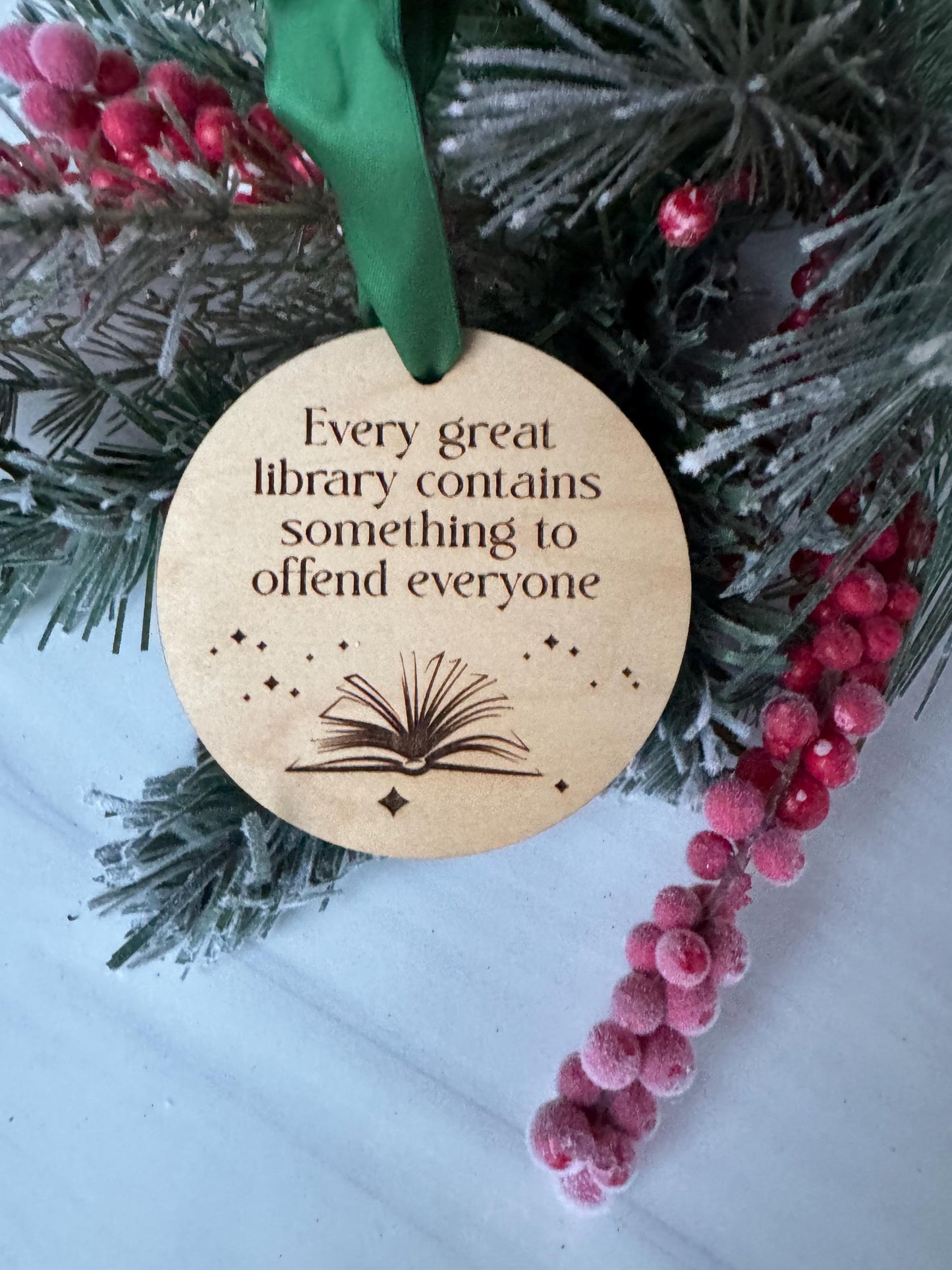 A wooden ornament with a book and the text &quot;Every great library contains something to offend everyone&quot; is displayed on a Christmas tree branch.