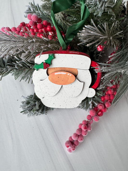 A white Christmas ornament with a red ribbon and holly leaves is displayed on a white surface, surrounded by red berries and a sprig of greenery.