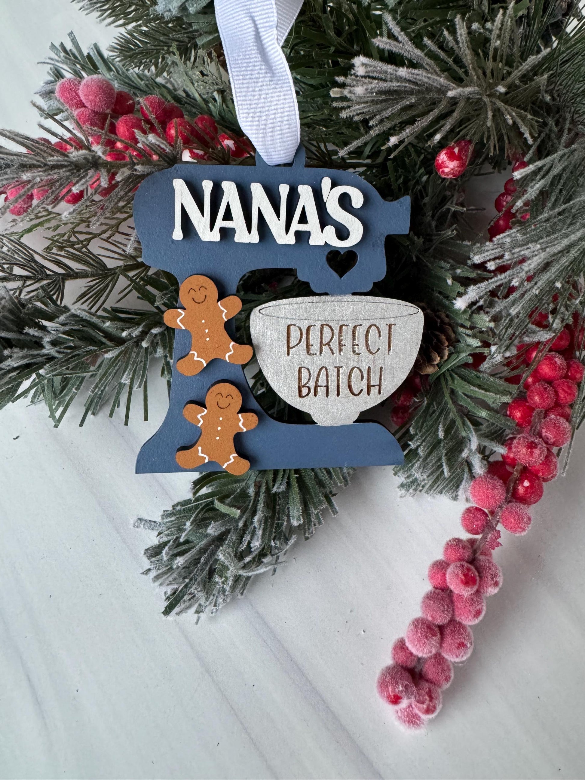 A Christmas ornament with the word &quot;NANA&#39;s&quot; written on it, surrounded by pine branches, red berries, and a white ribbon.