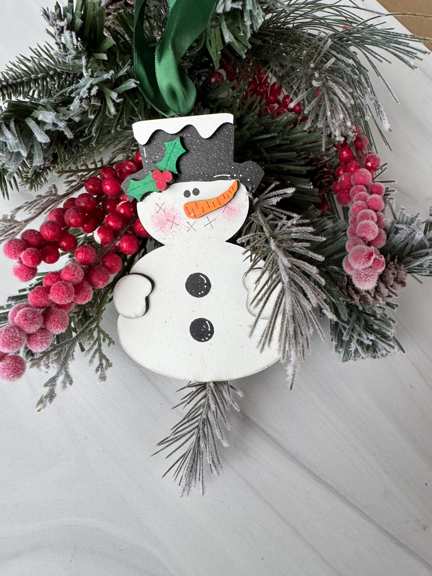 A festive wreath adorned with red berries, pine branches, and a snowman ornament is displayed on a white surface.