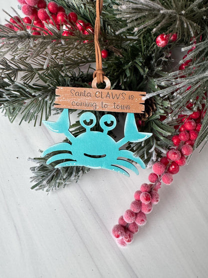 A Christmas ornament in the shape of a blue crab, with a sign that says "Santa's coming to town". The ornament is hanging on a pine branch with red berries, and the background is a white surface.
