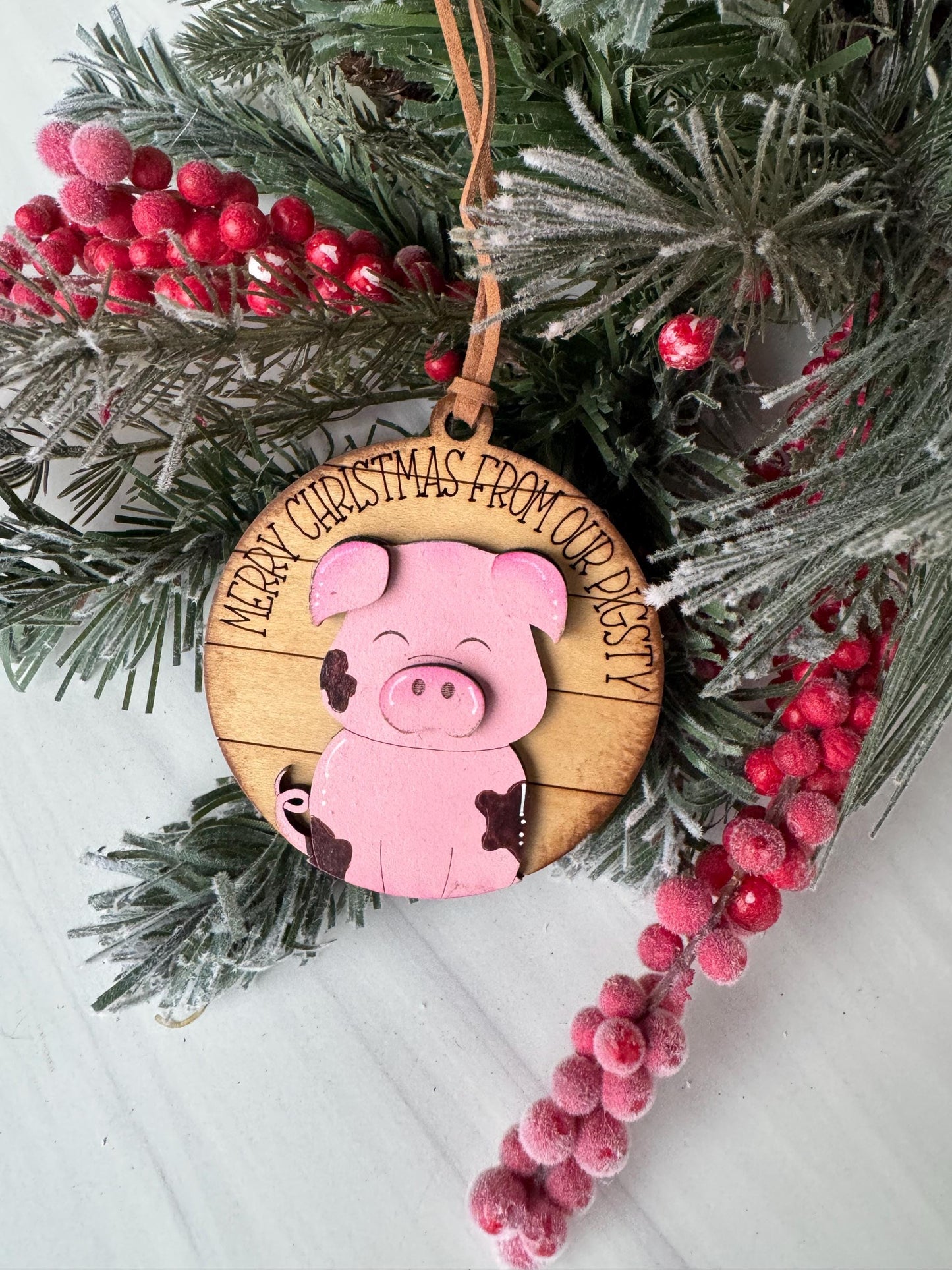 A Christmas ornament featuring a pink pig is displayed on a pine branch with red berries.