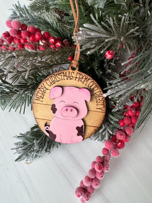 A Christmas ornament featuring a pink pig is displayed on a pine branch with red berries.