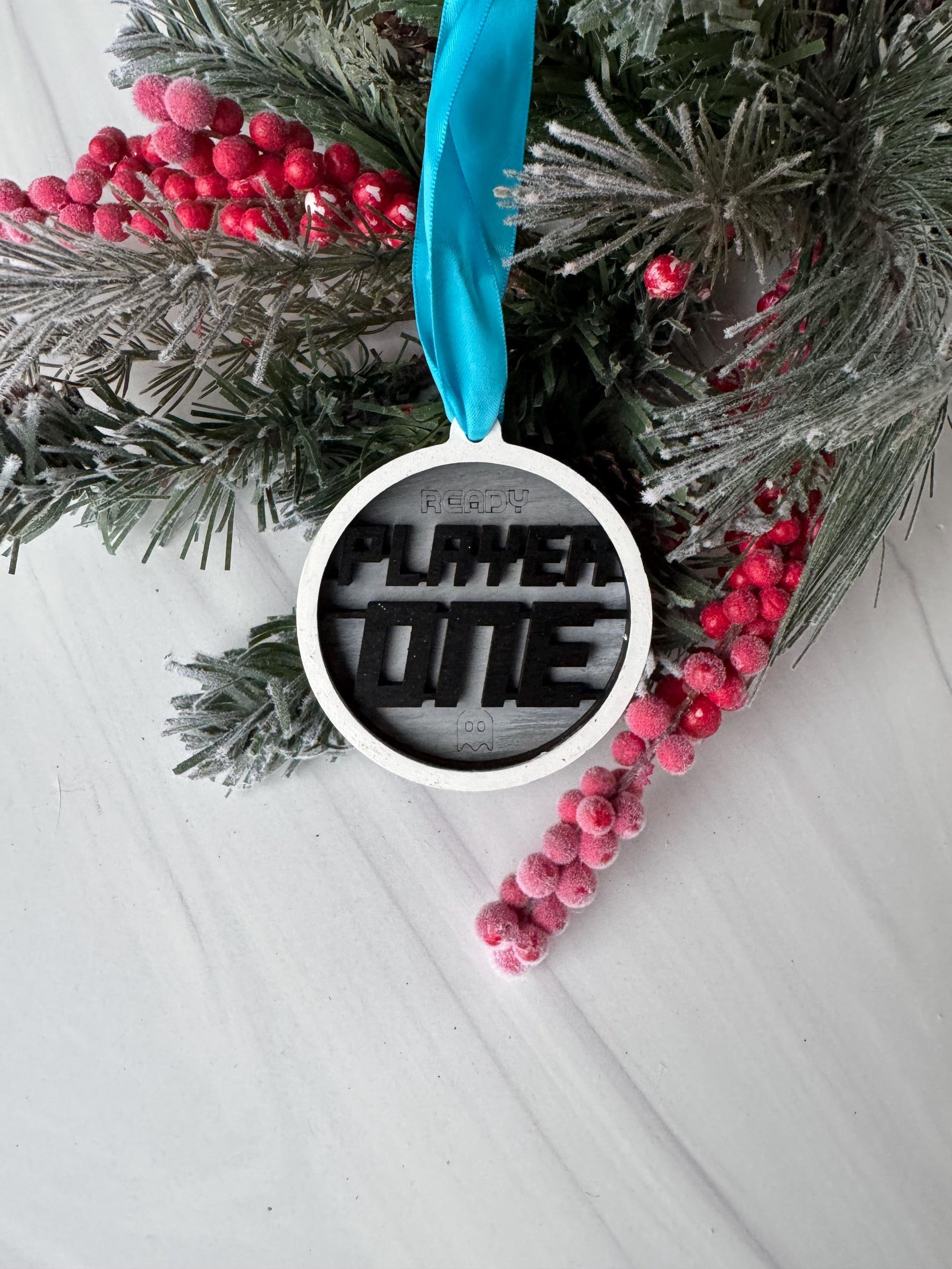 A Christmas ornament with the text &quot;FLY ME&quot; is displayed on a white surface, surrounded by pine branches, red berries, and a blue ribbon.